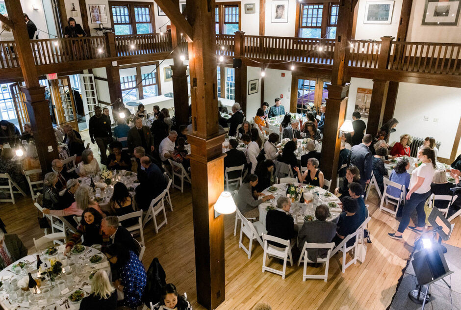 A brown wood room with tables set up, and wood columns, has guests sitting at the tables