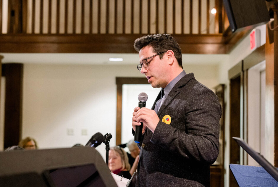 A man on stage speaks into a microphone while looking down at his notes