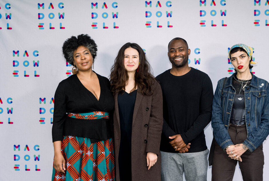 A group of four people smile and stand in front of a white and colorful MacDowell logo branded step and repeat