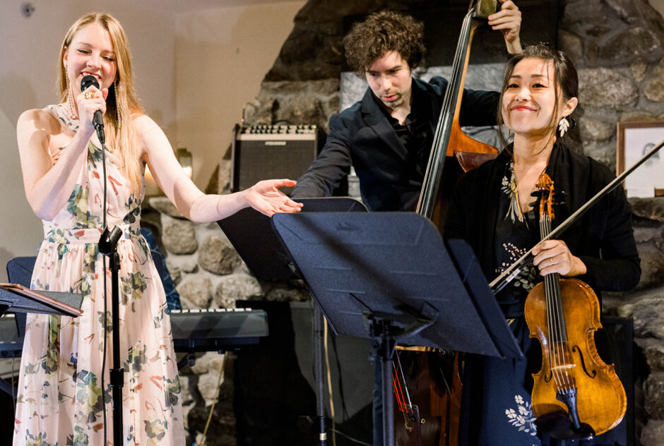 A woman in a long pink dress speaks into a microphone and gestures to a violinist and a bass player that are standing next to her