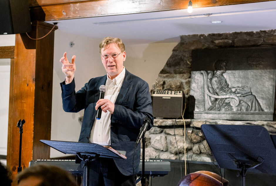 A man speaks into a microphone on stage and raises an imaginary glass in toast