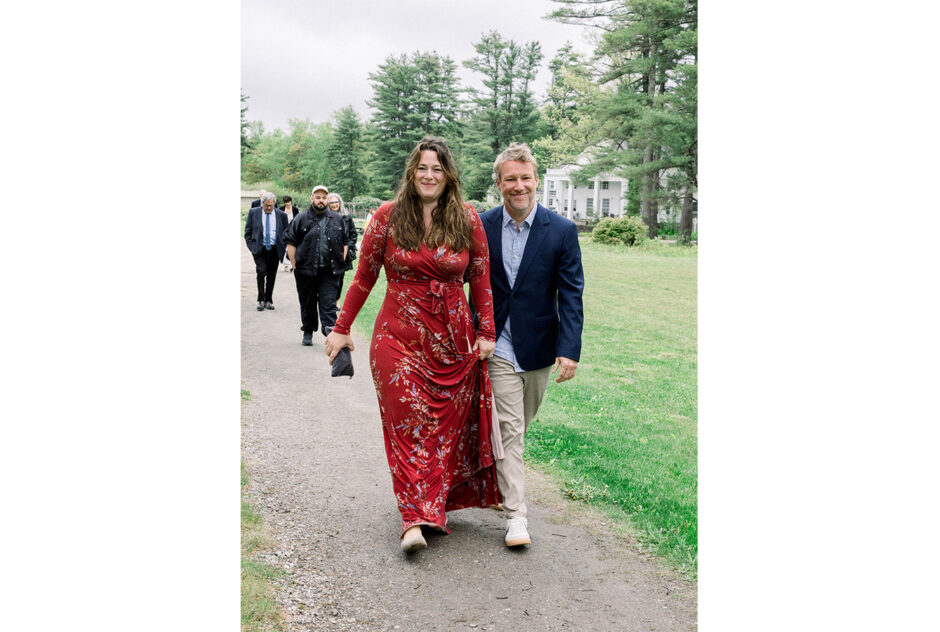 A woman in a red dress and a man in a blue blazer walk down a path lined with grass