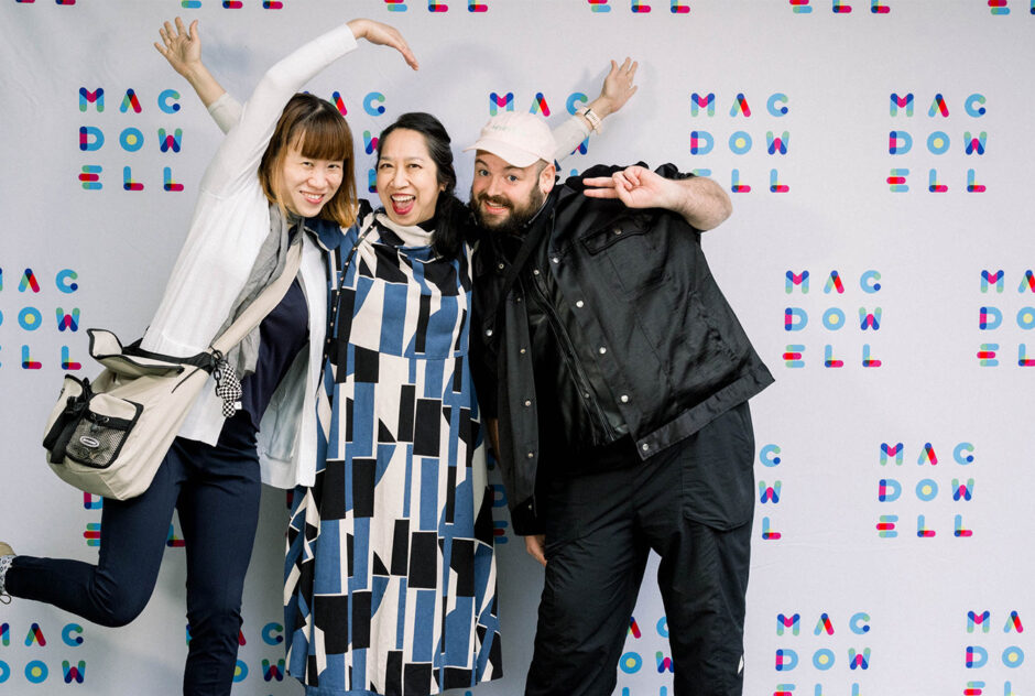 Three people smile and pose playfully in front of a white and colorful MacDowell branded step and repeat