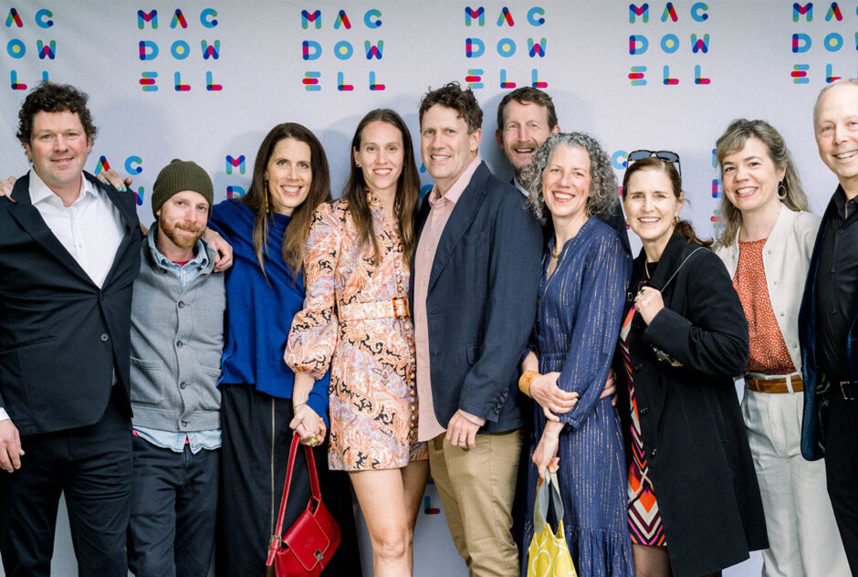 A photo of a group of people of a range of ages standing in front of a white and colorful step and repeat that says MacDowell