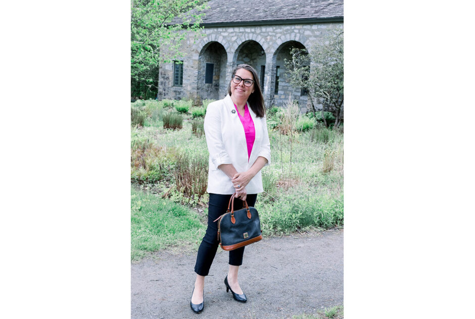 A woman wearing a white blazer and pink top poses and smiles for the camera in front of a grey stone building