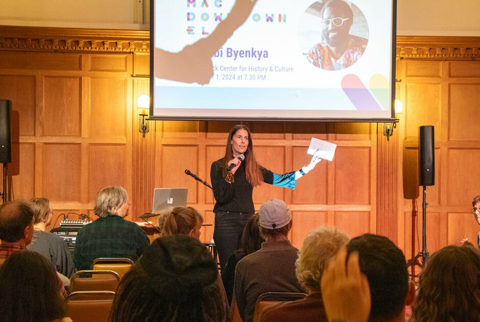 A woman stands, microphone in hand, at the front of a room. People sit in rows of chairs in front of her. She speaks to them while holding up a sheet of paper. Behind her is a projector screen.