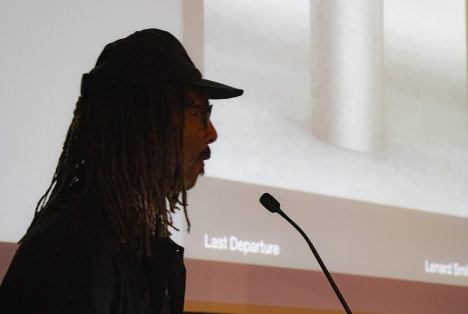 A man stands at a podium, presenting to an audience. Behind him, an image is projected onto a screen