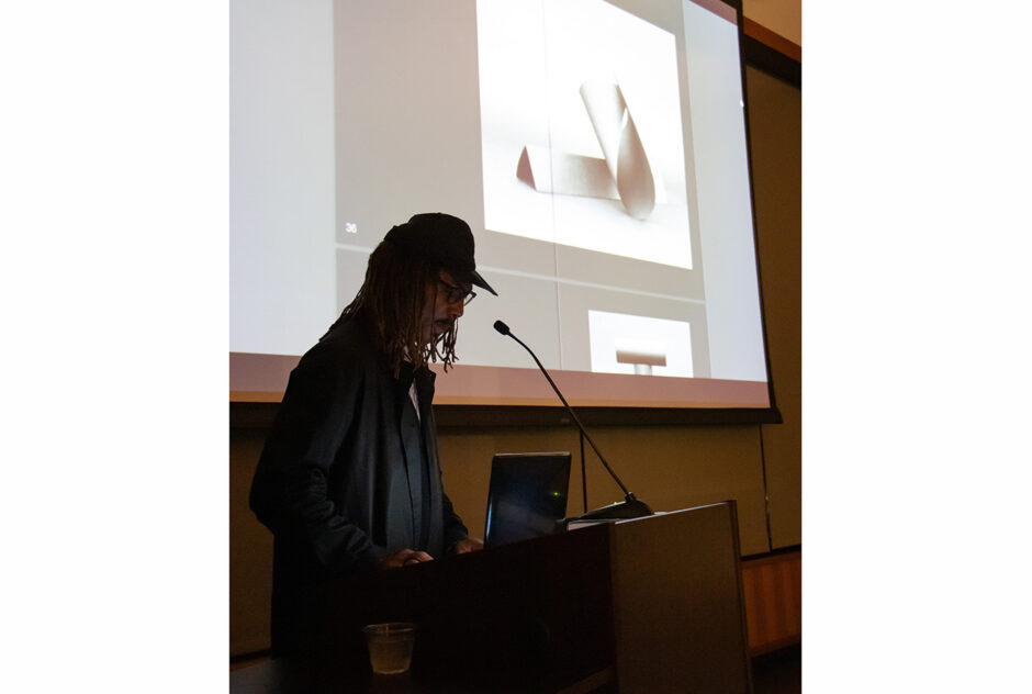 A man stands at a podium, presenting to an audience. Behind him, an image is projected onto a screen