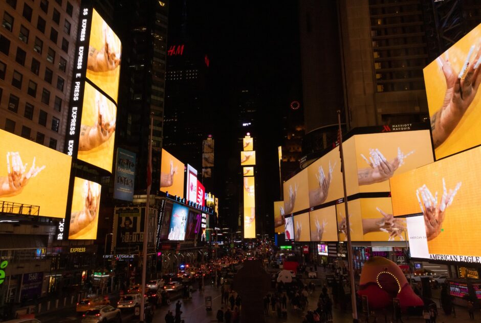 On multiple billboard screens above a busy city street by night, a still from a video art piece by Pamela Council showing an elaborately manicured hand is projected.