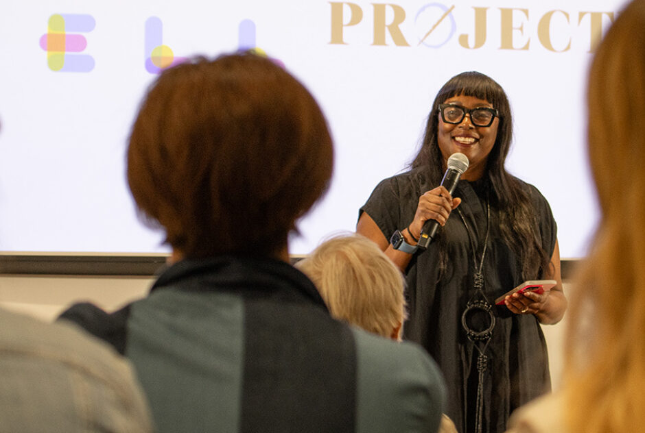 A Black, female-identifying person, Chiwoniso Kaitano, stands in a crowded room with a mic in her hand.