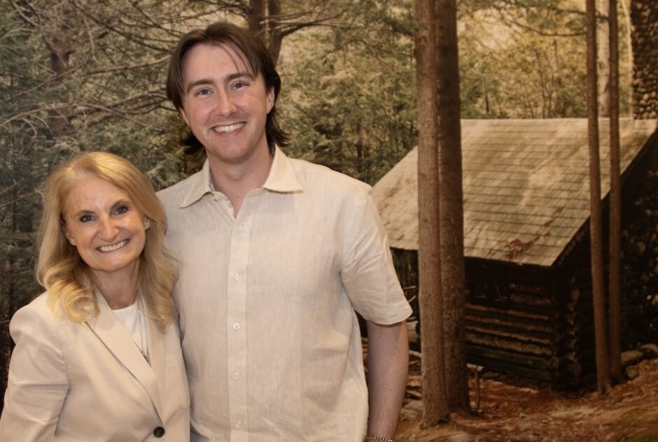 A female-identifying person (Christine Fisher) stands for a portrait with male-identifying person Emerson Sistare. Behind them is a mural of a cabin. They smile and look at the camera. 10. Three female-identifying people stand for a portrait in a crowded room. They smile and look at the camera.