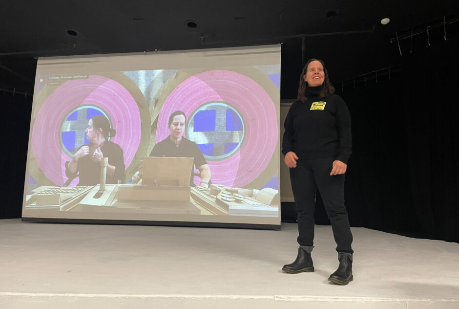 A woman, dressed in all black, stands on a stage. Behind her, a projector screen is displaying a clip from one of her videos.