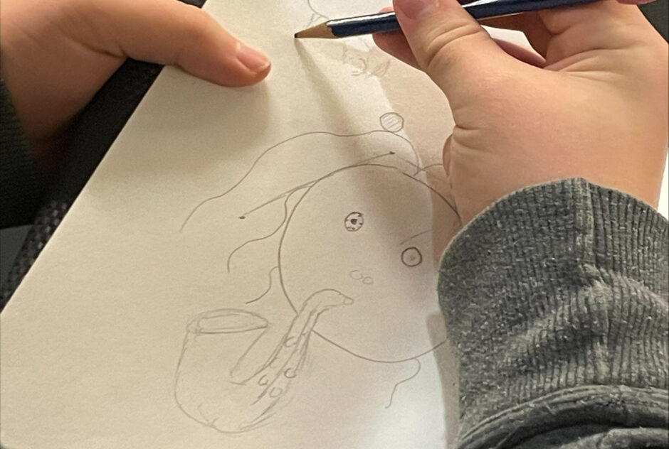 A close up image of a students hands as they use a pencil to draw a character on a sheet of paper.