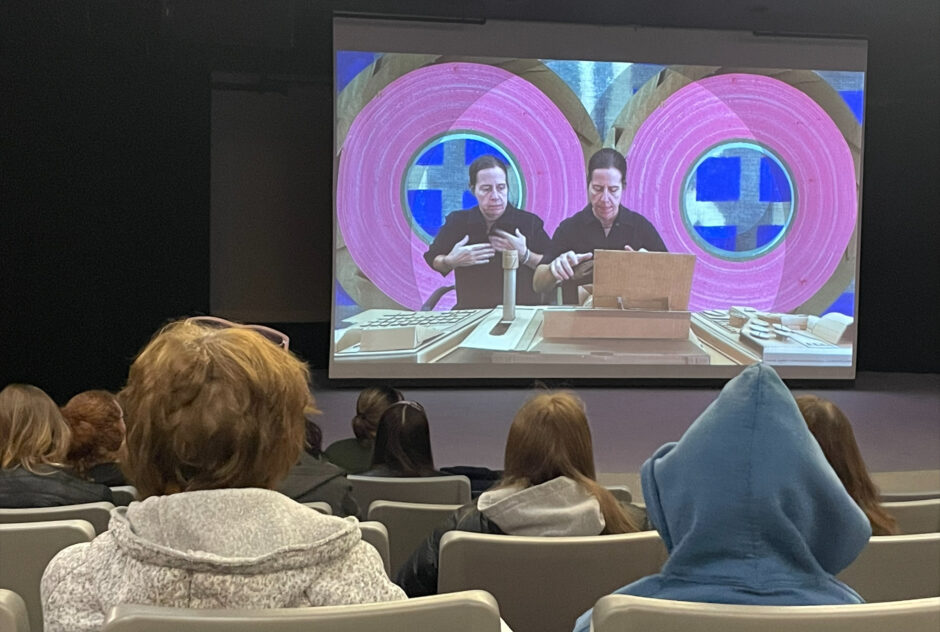 Students, sitting in rows of seating in a small auditorium, look toward the stage at the front of the room where a projector screen is displaying a short film.