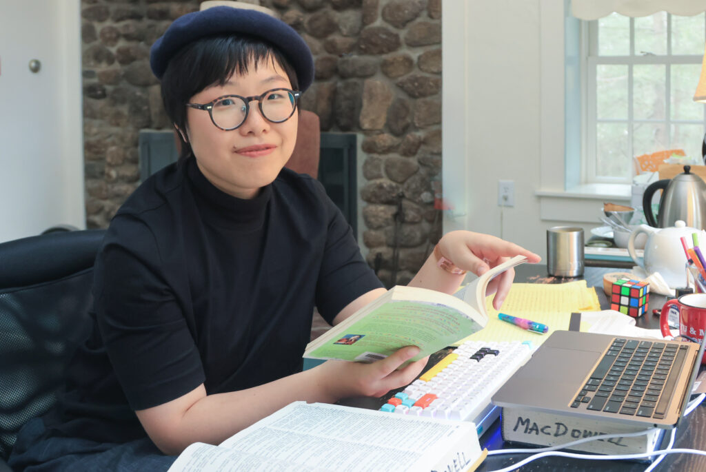 A person sitting at a desk holding a book in their hands. They are smiling and looking at the camera, papers are strewn about the desk.