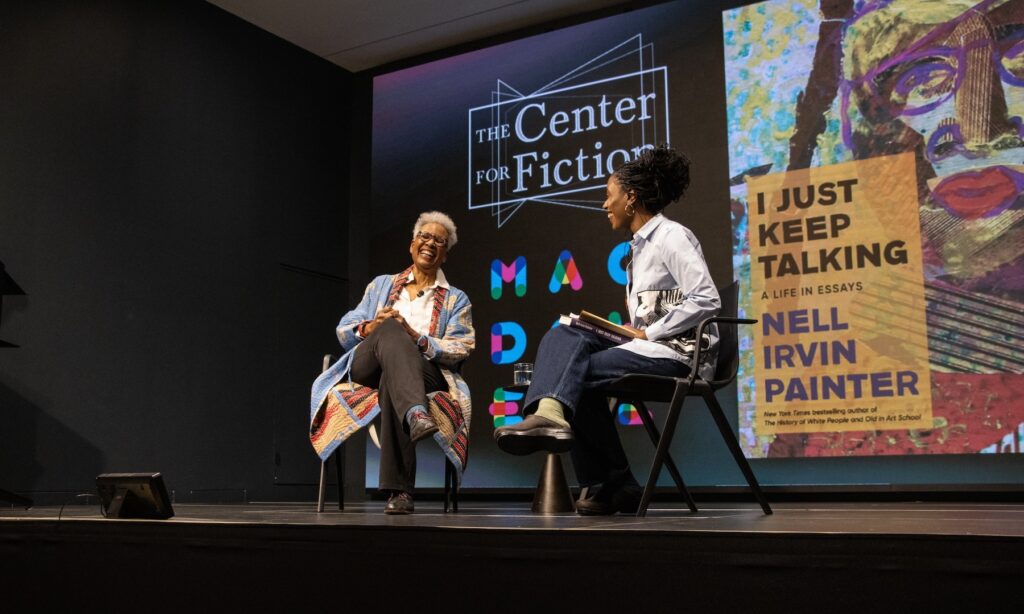 Two women seated in chairs on a stage laugh in the midst of a book discussion. Behind them, a projector screen displays the logos for the Center for Fiction and MacDowell and an image of the cover of Nell Painter's book, I Just Keep Talking.