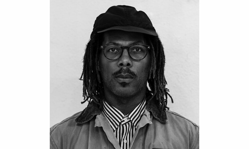 Portrait of a man wearing a black baseball cap, a striped button down shirt, and a jacket. He is looking straight into the camera, not smiling. The image is black and white