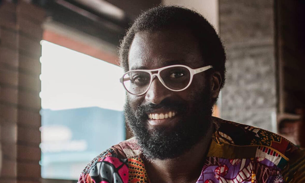 A smiling person wearing a colorfully patterned shirt and white rimmed glasses. The room is fairly dark and there is a large window in the background lighting the space