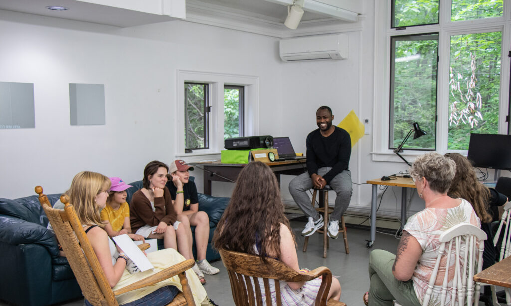 A group of students gather inside of a studio, sitting in chairs and on couches. They are listening to a presentation from a Fellow who is sitting on a stool next to a projector.