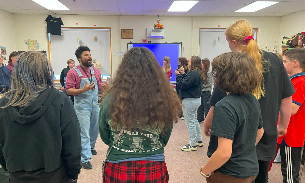 A man in overalls and a red long sleeve shirt stands in the center of a room, speaking and gesturing with his hands. A large group of students gather in a circle around him.