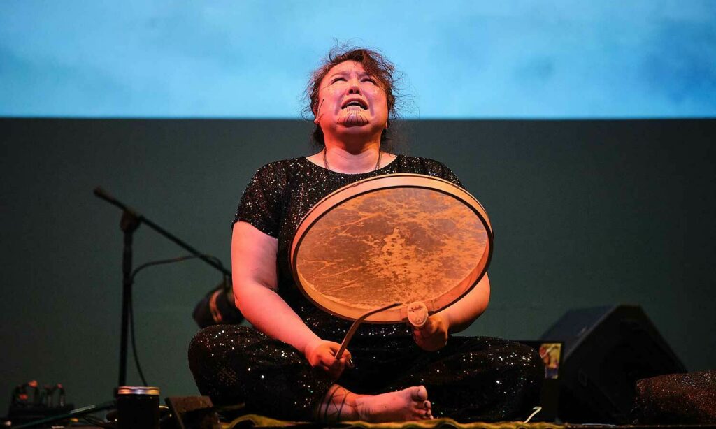 Allison Akootchook Warden is drumming and singing, cross-legged on stage during 2022's First Nations Performing Arts celebration at Portland Institute for Contemporary Art.