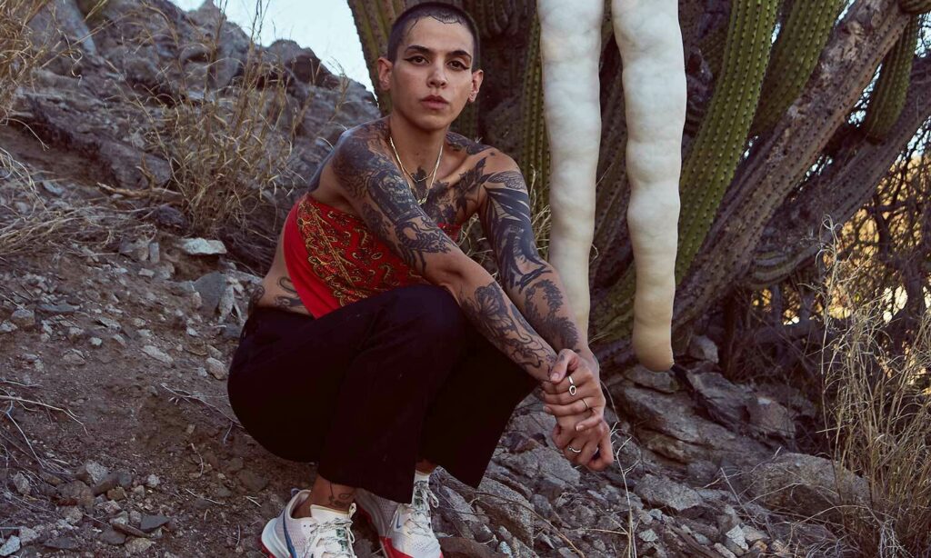 Ana Armengod squats with her tattooed arms extended over her knees wearing black pants. On the hillside beside her is a saguaro cactus from which hangs an apparent effigy.