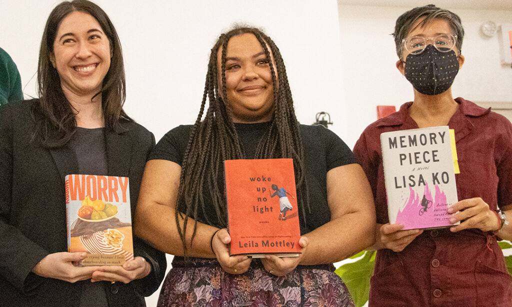 Three people stand shoulder to shoulder, smiling and holding books in front of them