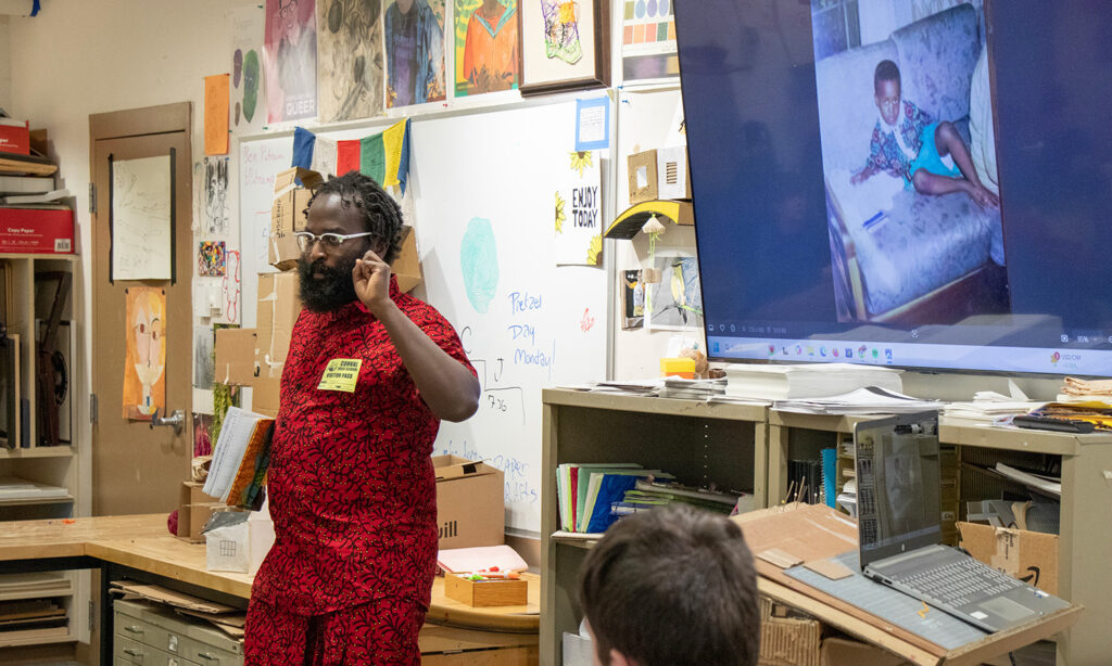 A person wearing a bright red outfit stands at the front of an art classroom, speaking and gesturing with their hand. The walls of the classroom are covered in artwork and papers. Behind the person is a large TV screen displaying an image of them from when they were a child.