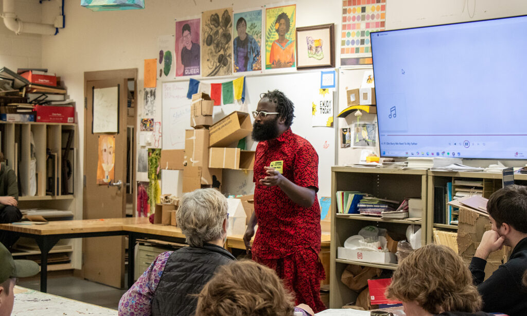 A man wearing a bright red outfit stands at the front of a classroom, gesturing with his hands as he speaks. The walls of the room are covered in papers, art, and art supply storage.