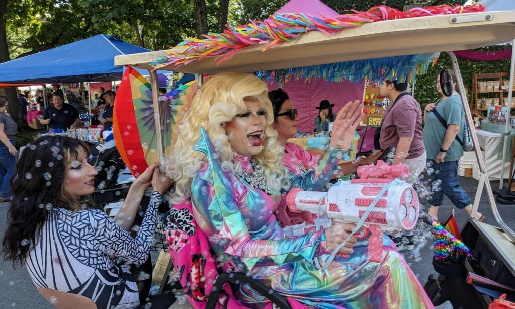 A golf cart with three immaculately dressed drag queens shoot bubbles from a bubble gun as they cruise around the festival