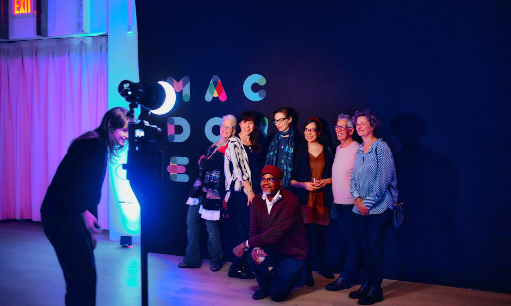 A group of smiling people pose for a picture in front of a black backdrop featuring the MacDowell logo.