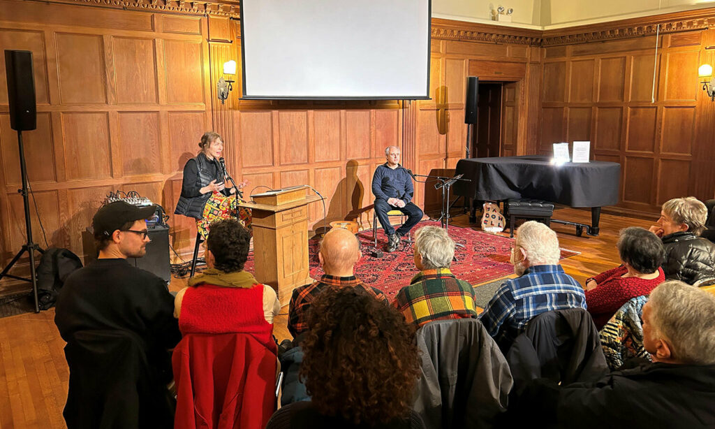 Two people sit at the front of a room, presenting to a crowd seated in rows of chairs. The room has warm lighting and is entirely paneled in wood