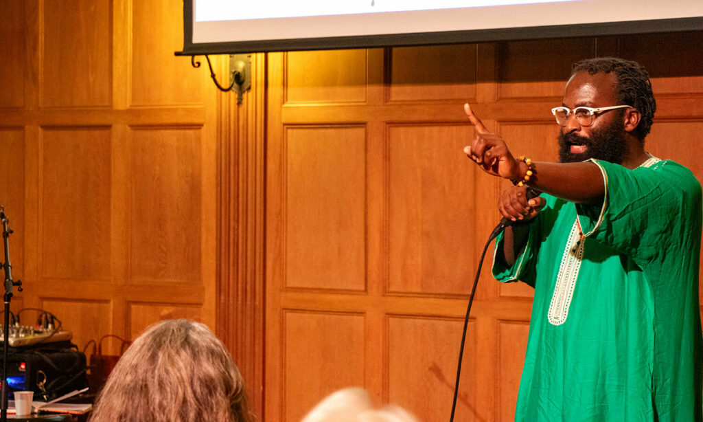 A man wearing abright green robe and white-rimmed glasses stands at the front of a room. While speaking into a microphone, he holds one hand up, pointing a finger.