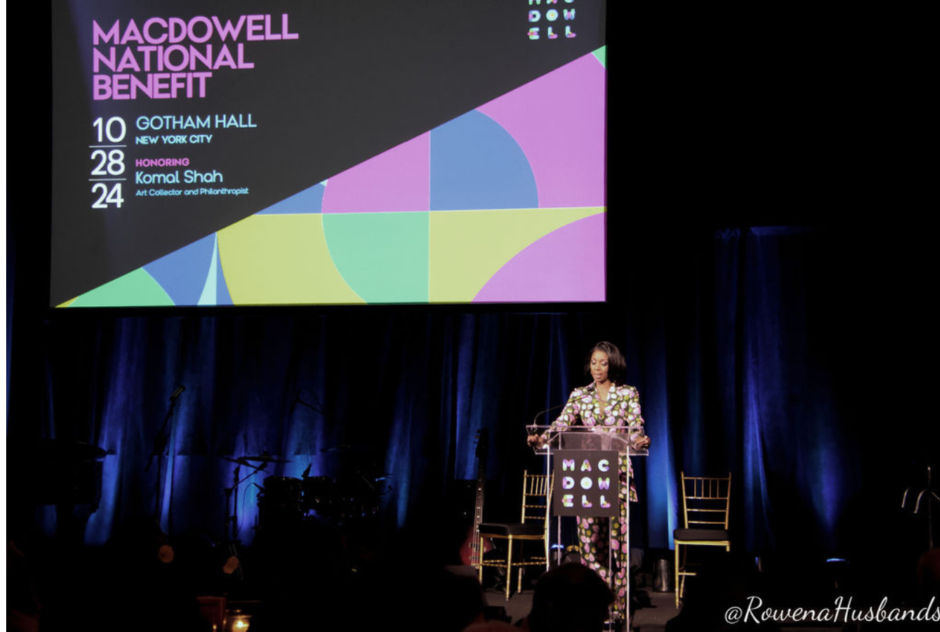 A woman stands on a podium on a stage in front of a colorful screen that says MacDowell National Benefit and a blue curtain