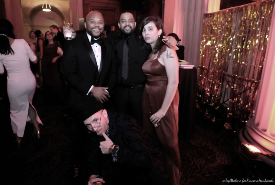 Three people wearing festive formal wear stand and pose for a photo, with someone posing in a squatting position beneath them, at a party