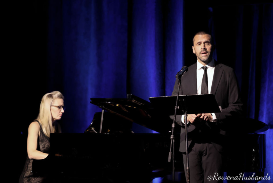 A blonde woman plays a piano onstage while a man in a suit sings