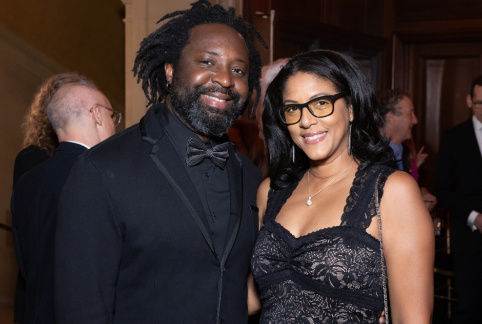 A person wearing a black suit and a person wearing a black lace dress and glasses smile for a photo at a party