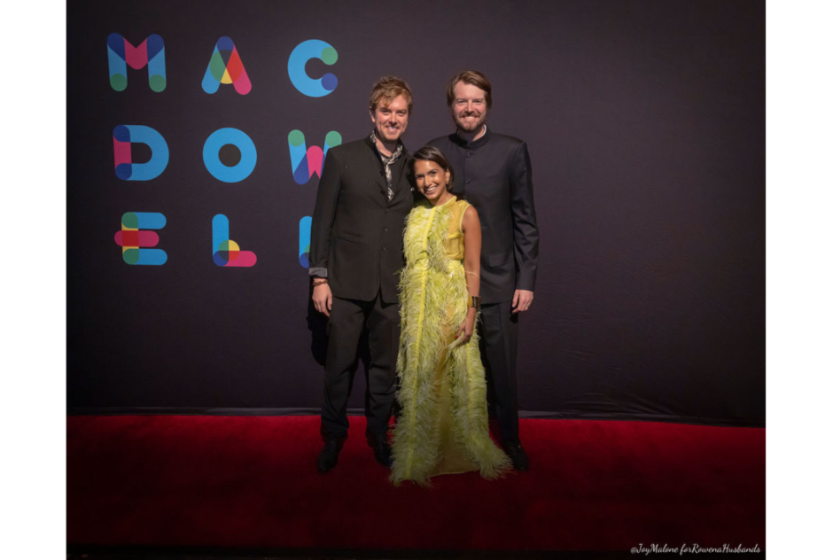 Two men in black suits and a woman in a yellow long dress smile in front of a black step and repeat