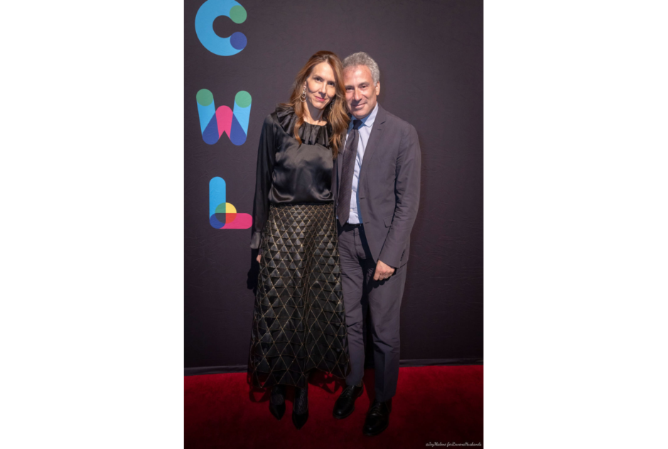 A woman in a black dress and man in a grey suit pose in front of a black step and repeat