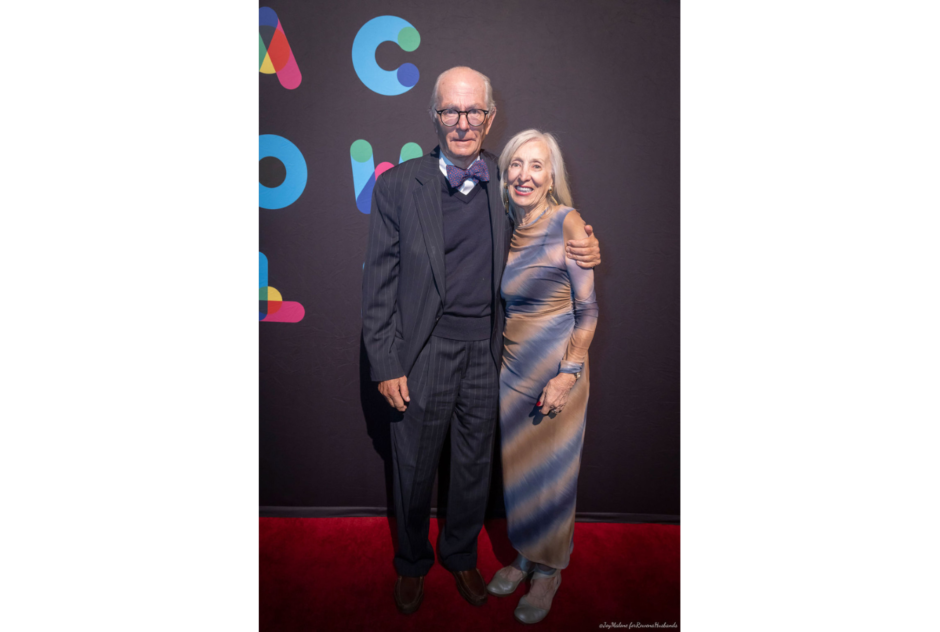 Two older white people in formal wear pose in front of a black step and repeat