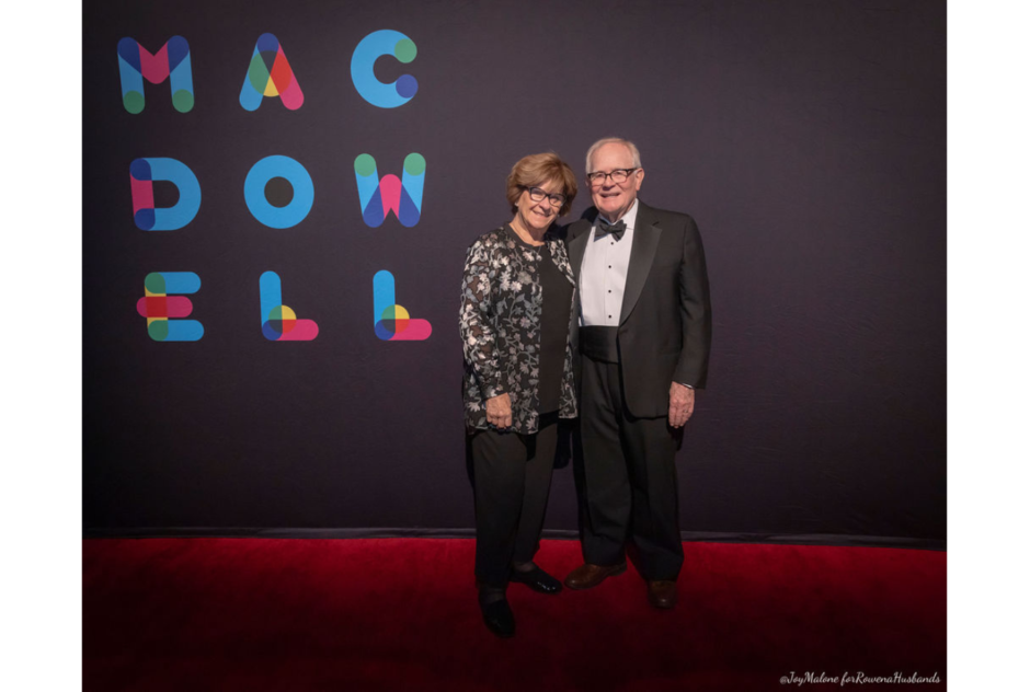 Two older white people in formal wear pose for a photo in front of a black step and repeat