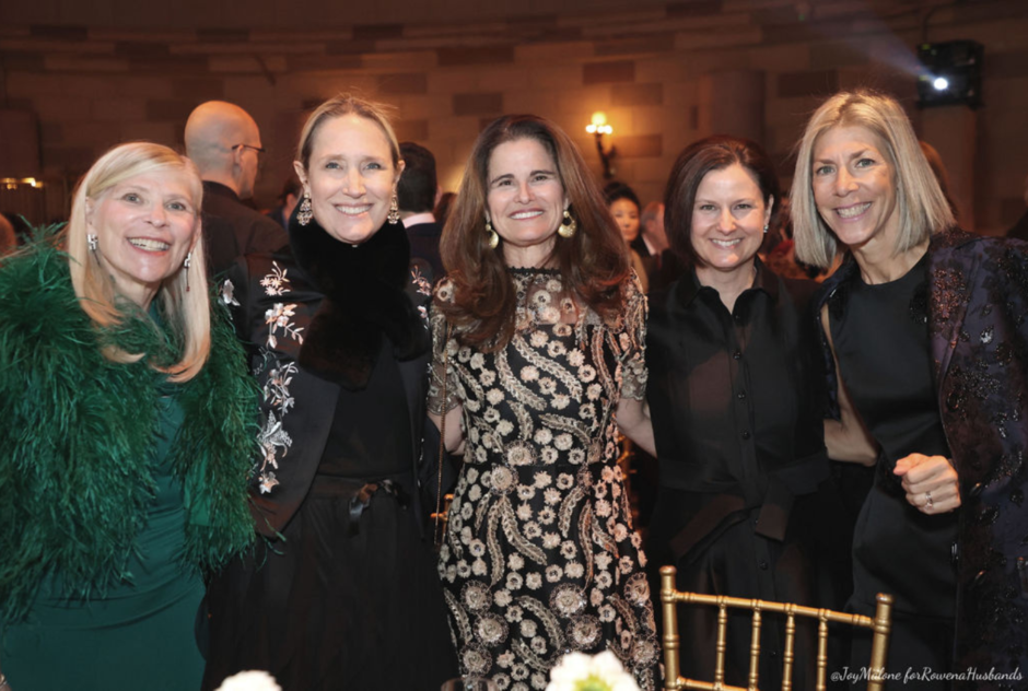 A group of white women wearing formal wear stand behind a dinner table at an event and pose for a photo