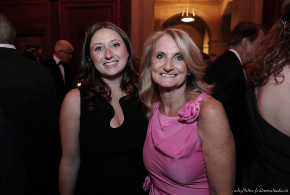 A young woman in a black dress and a middle aged woman in a pink dress smile for a photo at a party