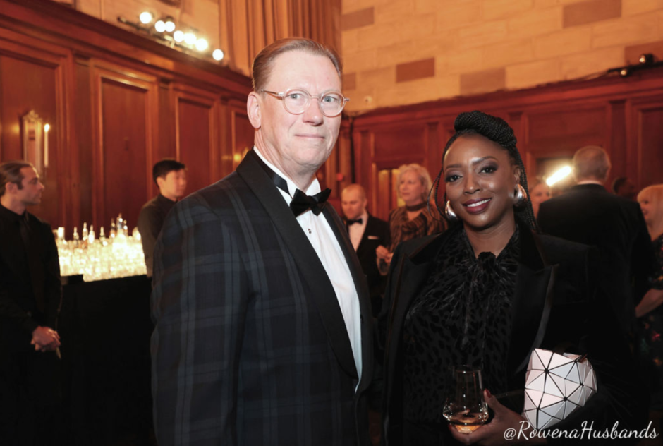 A man in a tuxedo and a woman in a black suit smile for a photo at a formal event