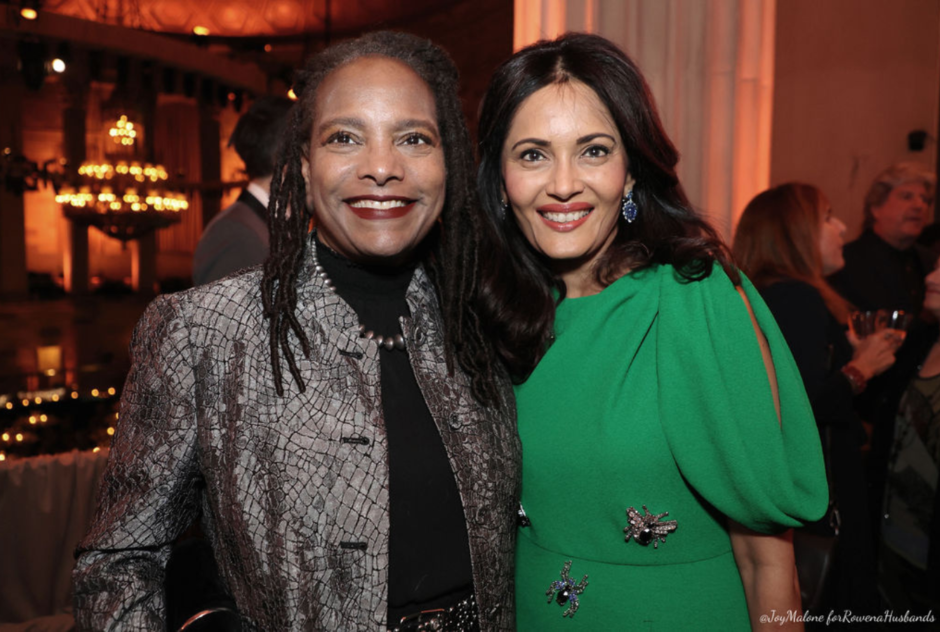 A woman in a metallic gray jacket and a woman in a green dress smile and pose for a photo