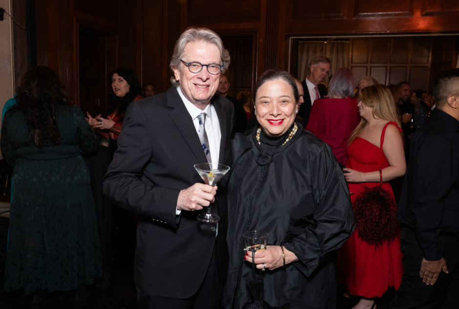 A woman and a man in black formal wear hold drinks and pose for a photo