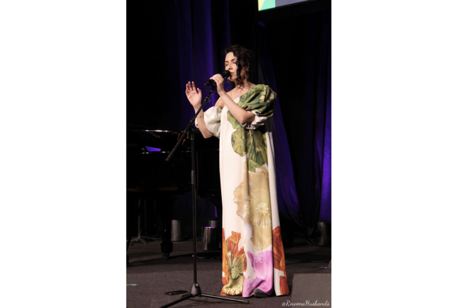 A woman in a long white and floral dress sings into a microphone on stage