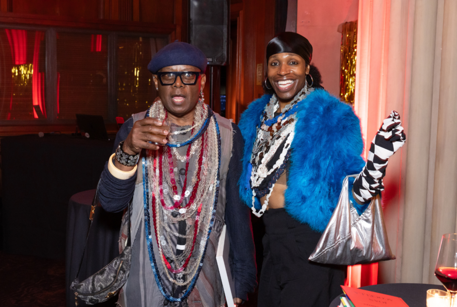 Two people in colorful outfits with sequined necklaces smile and hold drinks in their hands