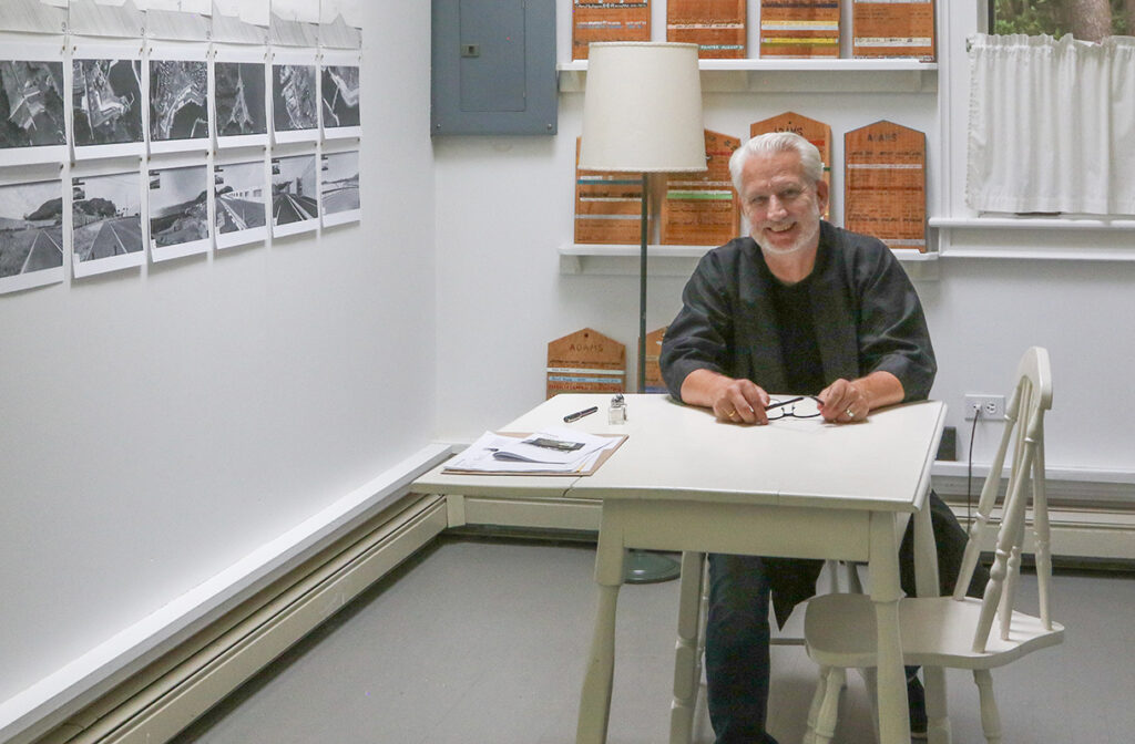 A gray-haired, bearded person sits at a white table in a white-walled artist studio. On the wall to his left are architectural renderings.