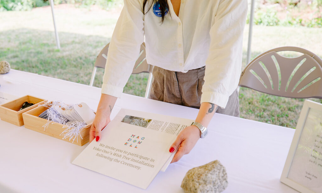 A woman stands at a long table and holds a small instructional sign. The table is laid with white cloth and materials for the Wish Tree are set out in bamboo boxes.